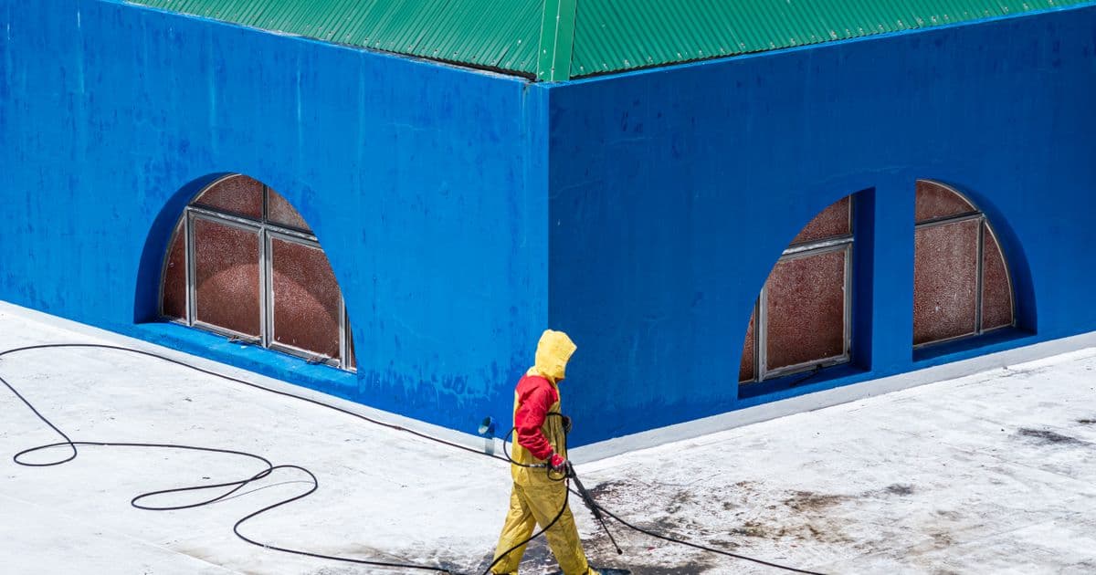 A worker pressure washing the exterior wall and walkway of a building on a sunny day